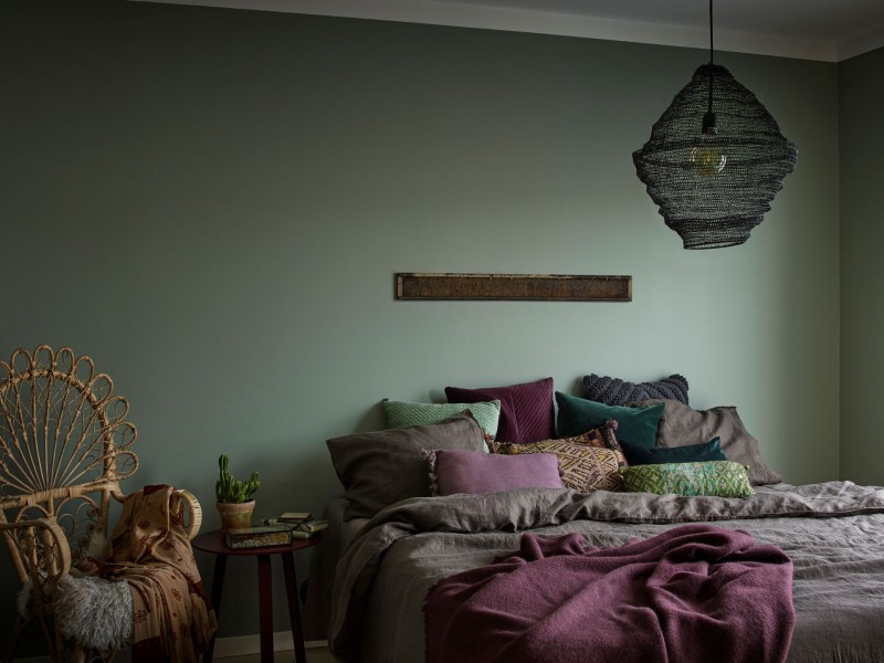 žalios spalvos akcentinė siena miegamajame su bohemiško stiliaus pagalvėmis ir lempa